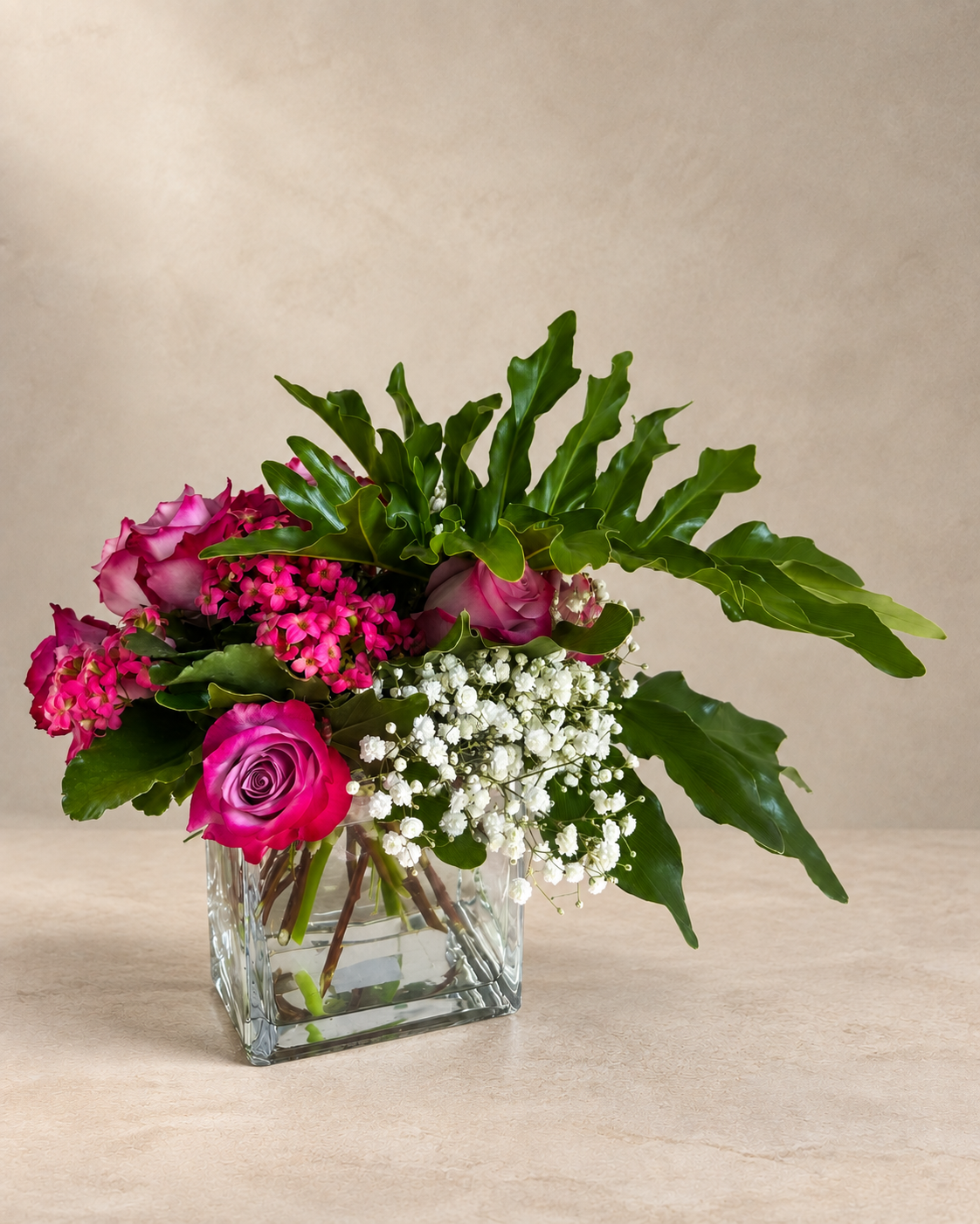 A vibrant floral arrangement featuring bright pink roses, magenta hydrangeas, green foliage, and white baby's breath in a clear square glass vase, set against a neutral beige background.
