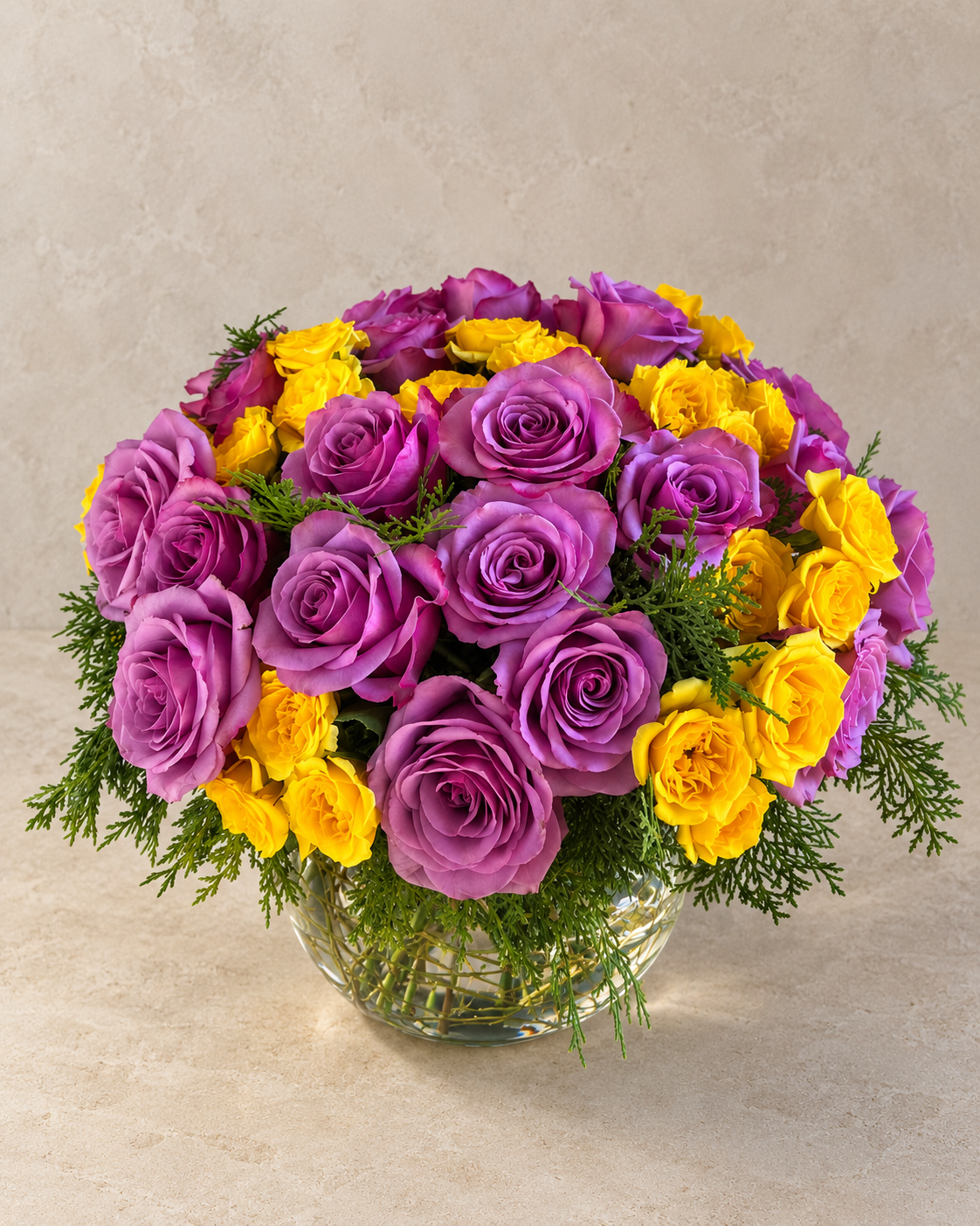 Elevated view of lavender and bright-yellow roses surrounded by cedar-like greenery in a transparent round glass vase.