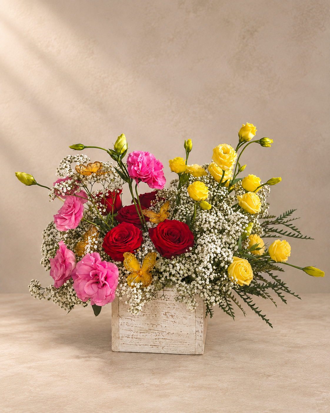 A vibrant floral arrangement featuring pink carnations, red roses, yellow roses, and white baby's breath in a distressed white rectangular wooden box, set against a warm beige textured background.