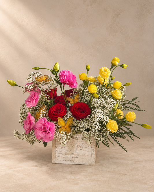 A vibrant floral arrangement featuring pink carnations, red roses, yellow roses, and white baby's breath in a distressed white rectangular wooden box, set against a warm beige textured background.