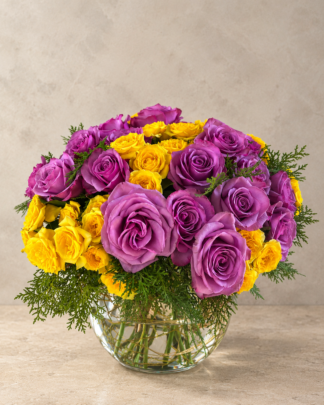 A vibrant flower bouquet featuring purple and yellow roses with green foliage, arranged in a clear glass vase on a light beige surface against a textured beige background.