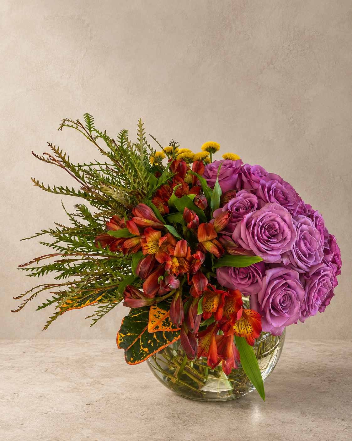 A vibrant floral arrangement featuring purple roses, red alstroemeria, and green foliage in a clear glass bowl vase, set against a neutral textured beige background.