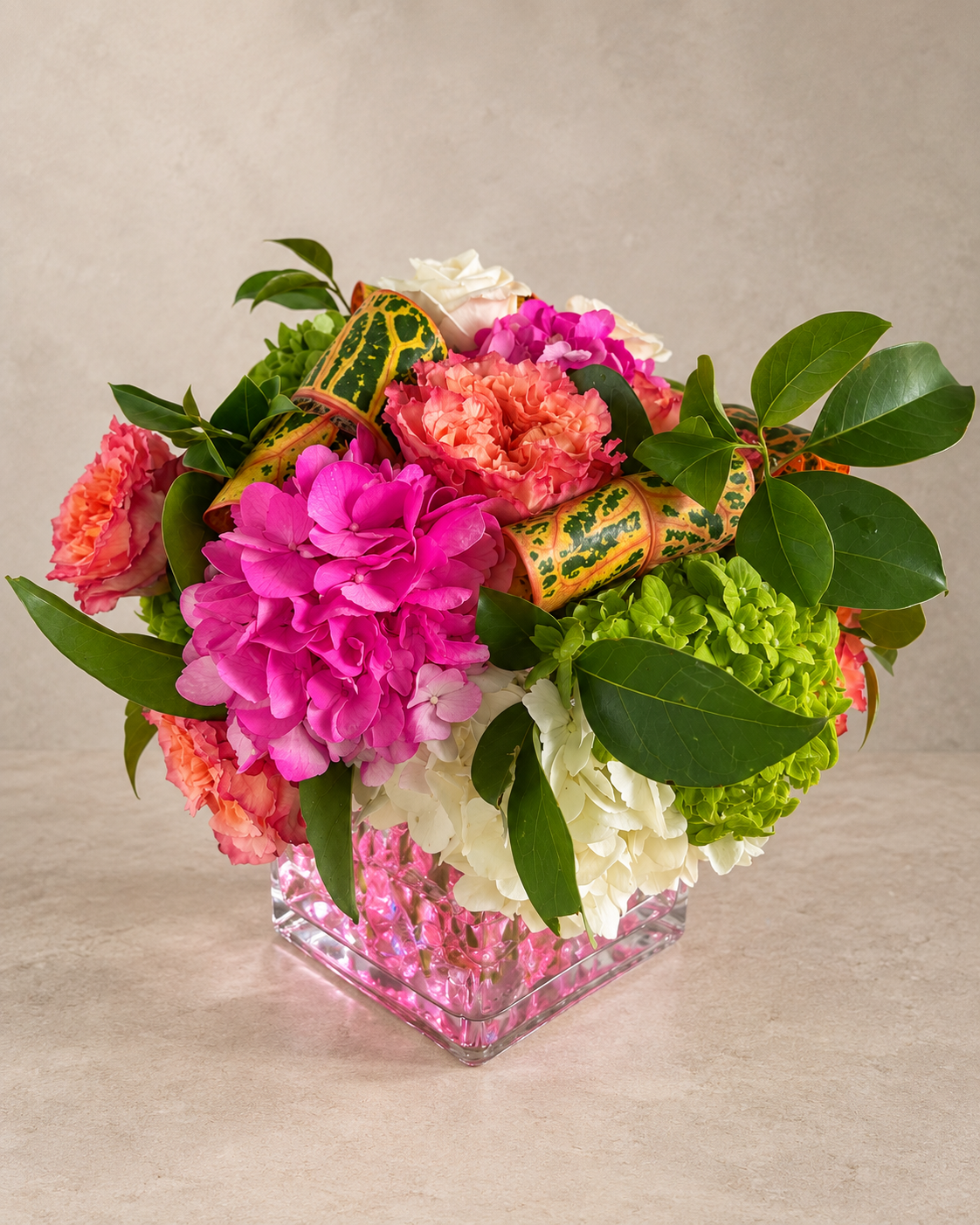 Reverse view featuring magenta, white and green hydrangea, coral ruffled blooms, croton leaves and glossy greenery above a pink-filled square glass vase.
