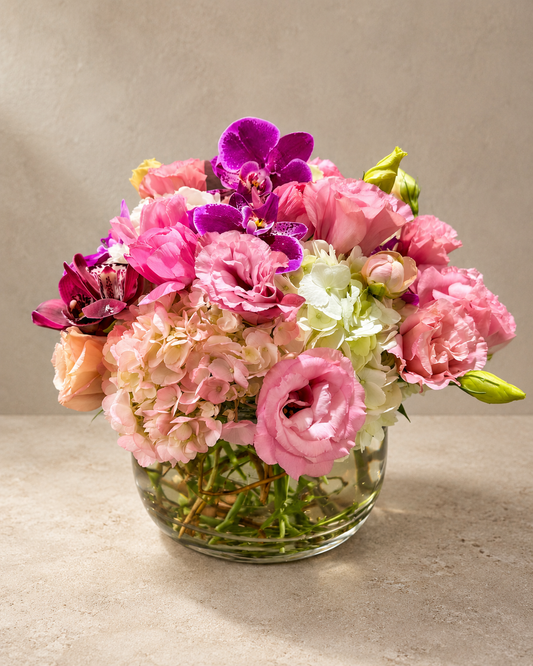 Pink Cocoon arrangement with pink lisianthus, hydrangea and purple orchids in a clear round glass vase.