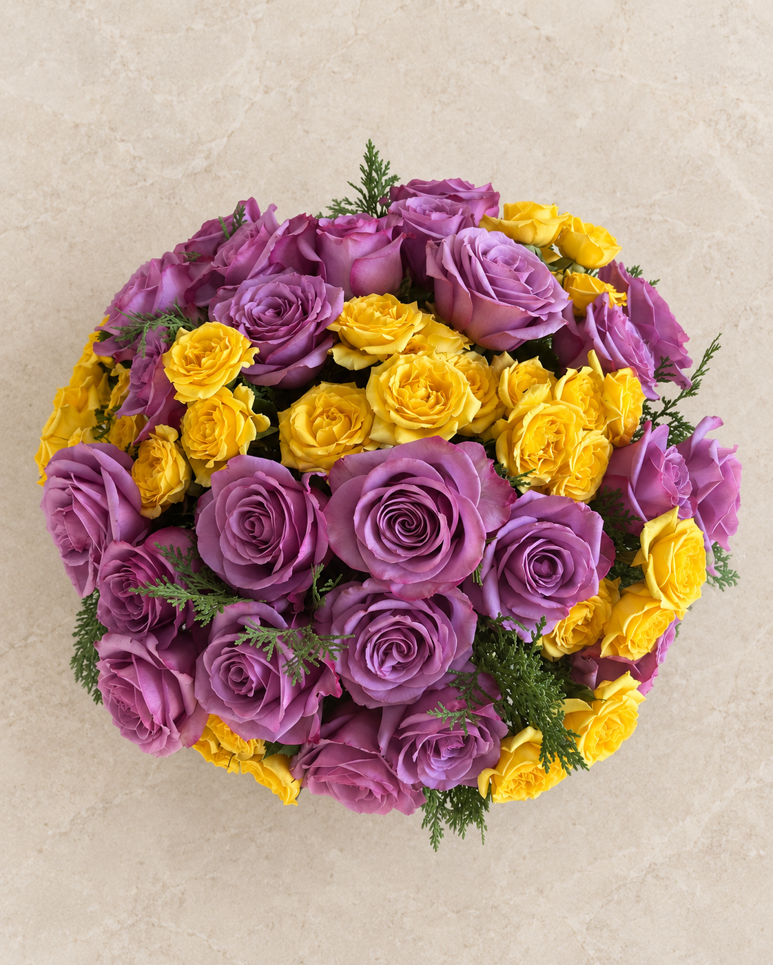 A round floral arrangement of purple and yellow roses with green foliage, photographed from above on a light beige textured background.