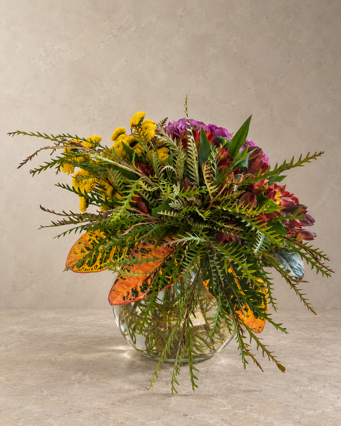 A vibrant autumn floral arrangement in a clear glass vase, featuring yellow ditsy flowers, purple protea blooms, green ferns, and orange-brown leaves, set against a neutral textured beige background.