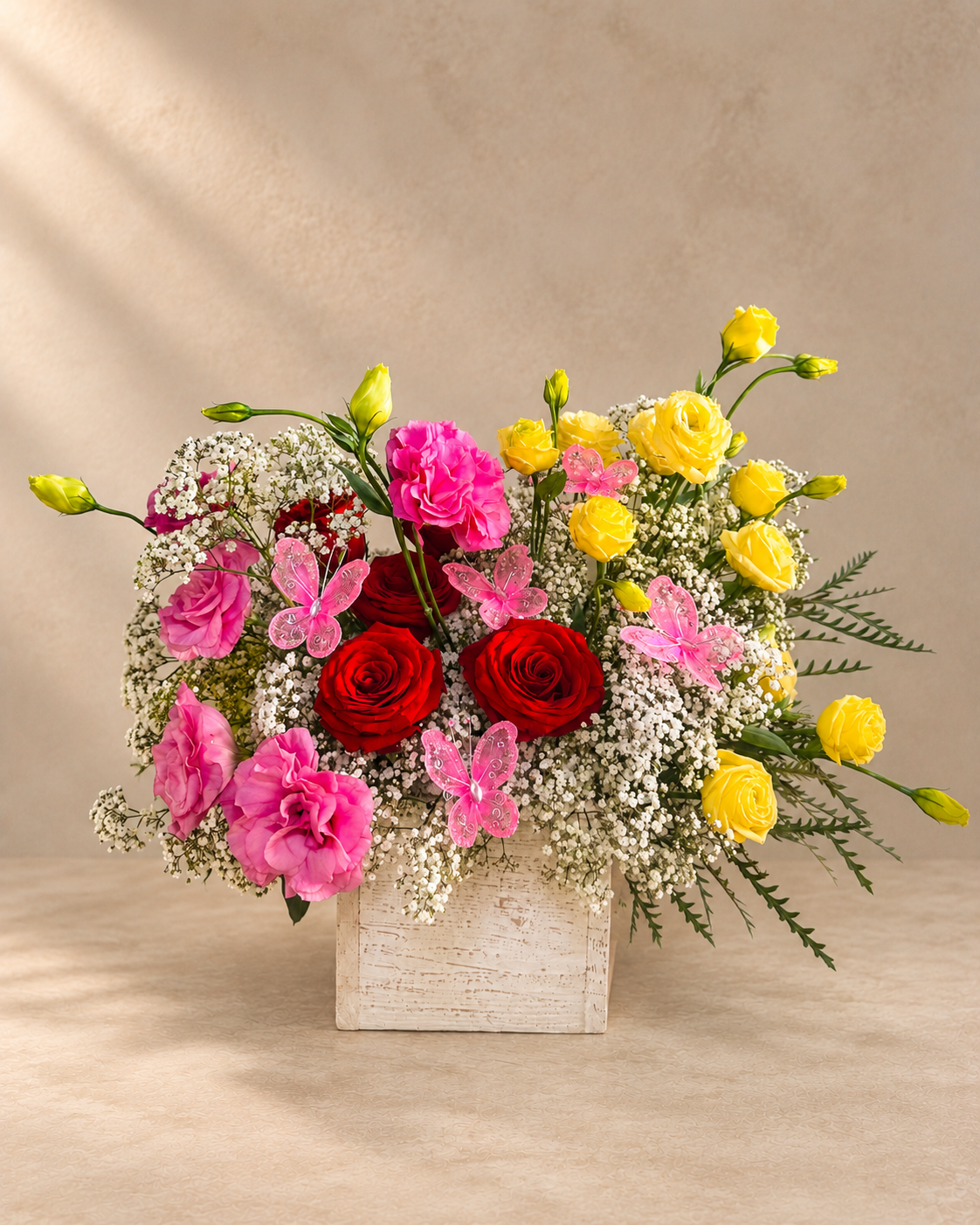 A vibrant floral arrangement in a rustic white wooden box, featuring red and pink roses, yellow carnations, pink carnations, butterfly flowers, and white baby's breath, set against a warm beige background.