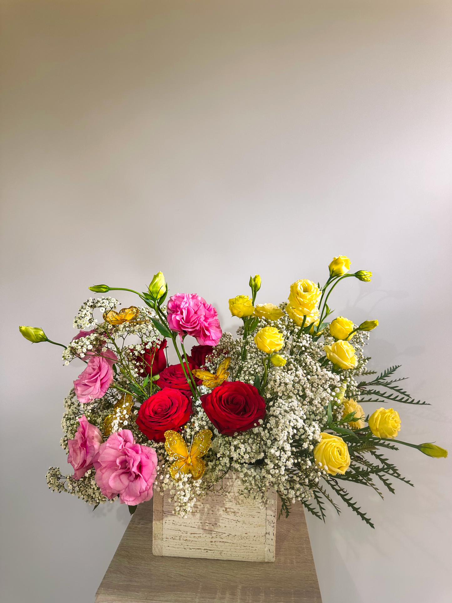 Elegant flower arrangement in a rustic square vase featuring vibrant red roses, pink lisianthus, yellow ranunculus, and delicate baby's breath. Accented with yellow and pink butterfly decorations, creating a romantic and whimsical design.