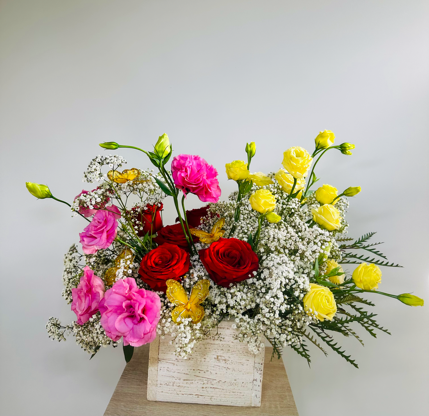 Fresh and colorful floral arrangement with red roses, pink lisianthus, and yellow ranunculus nestled in baby's breath and greenery, displayed in a wooden box. Yellow butterflies add a playful touch to this beautiful display.