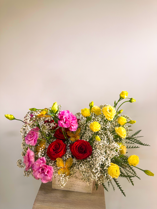 Charming floral arrangement in a textured wooden vase, showcasing red roses, pink lisianthus, yellow ranunculus, and baby's breath. A soft and romantic design complemented by greenery and butterfly accents.