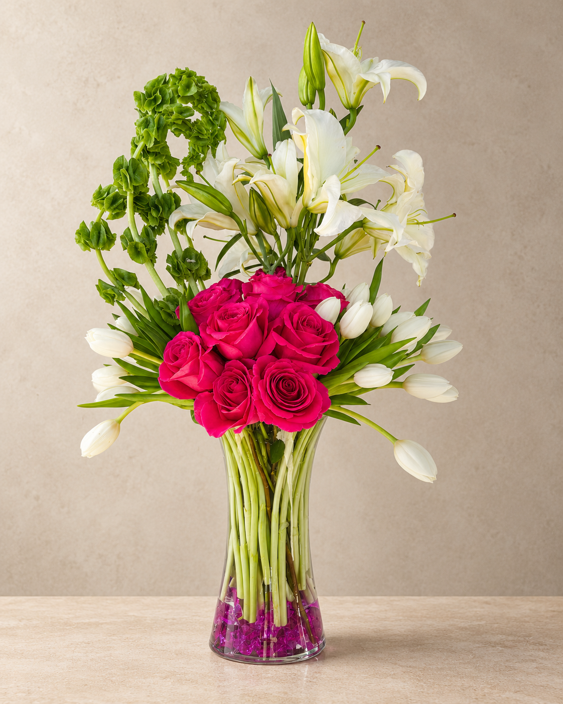 A floral arrangement in a clear glass vase on a light stone surface against a warm beige wall. The bouquet includes bright pink roses, white lilies, white tulips, and green foliage, with purple filler flowers at the base of the vase.
