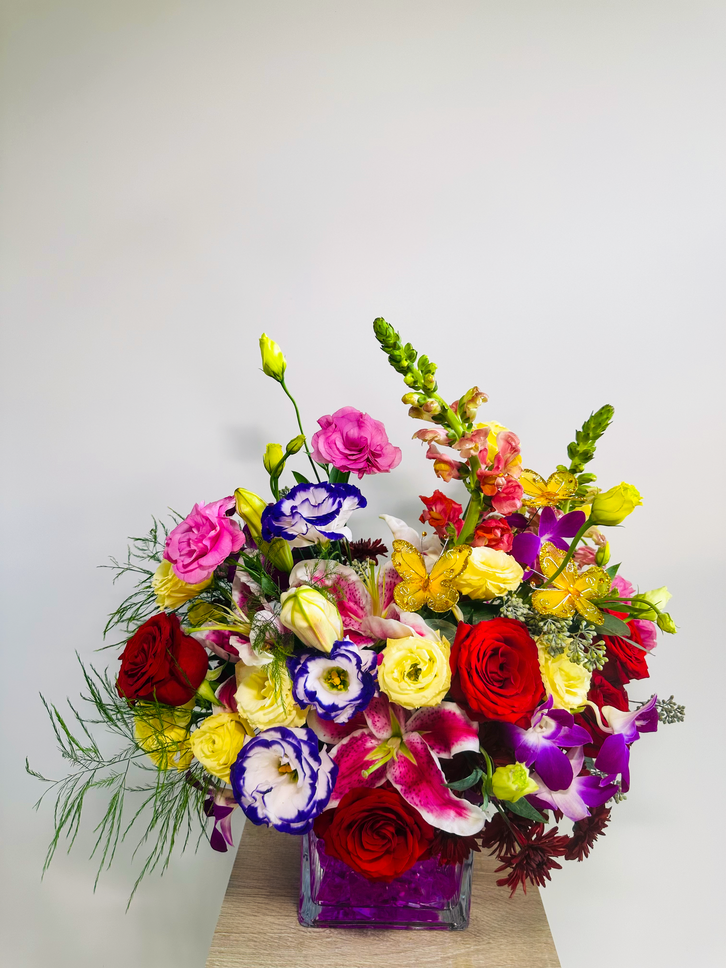 Vibrant floral arrangement featuring red roses, pink lilies, yellow snapdragons, and purple lisianthus in a clear glass vase with pink gems, perfect for Houston events.