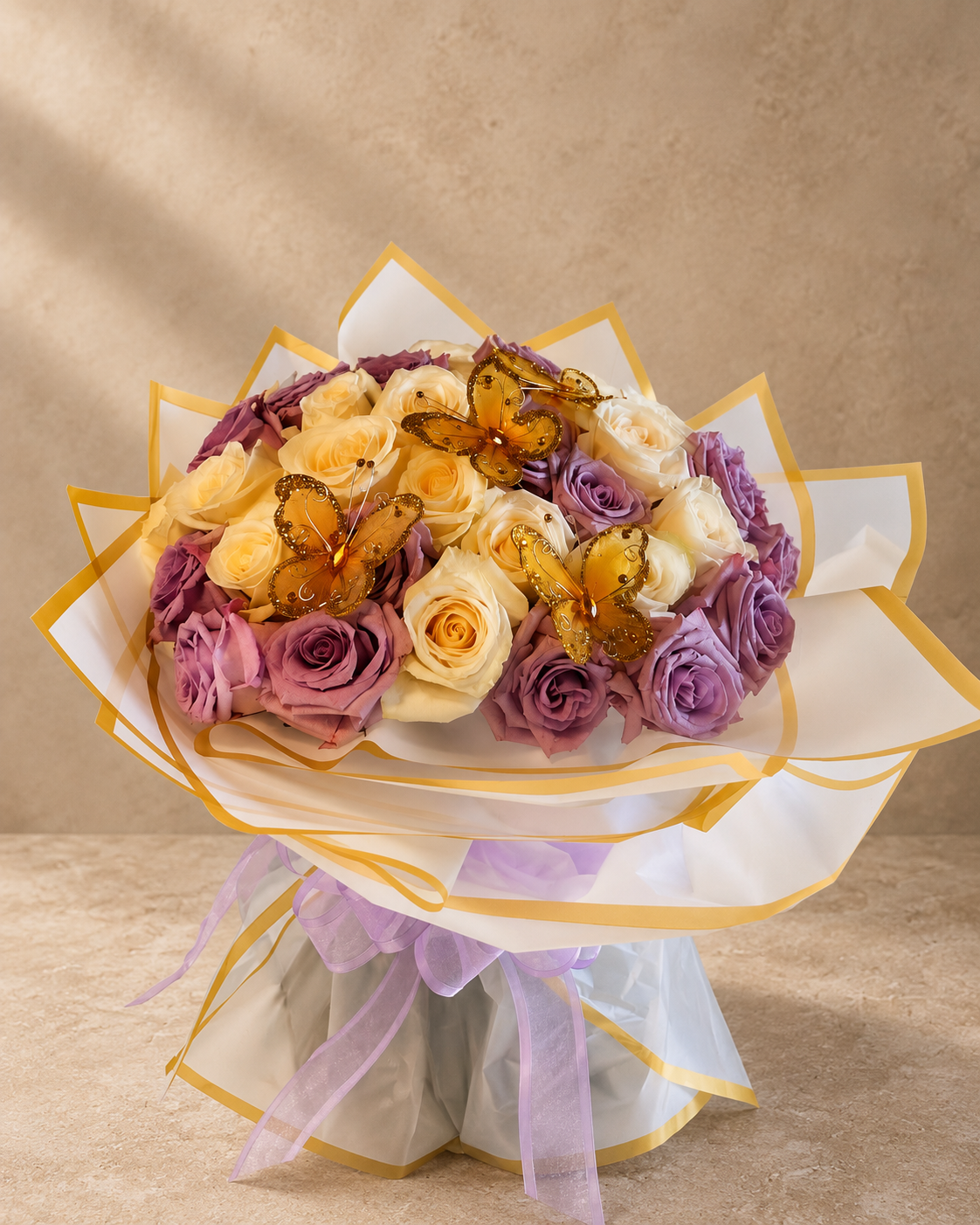 A bouquet of purple and cream roses wrapped in white paper with gold trim and a purple ribbon, decorated with three amber butterflies, set against a neutral beige background.