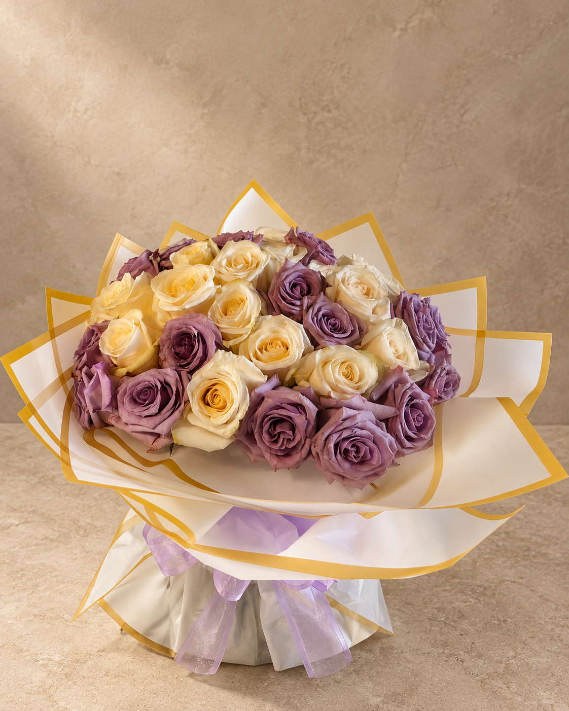 A bouquet of mixed white and purple roses wrapped in white paper with gold trim and a sheer lavender ribbon, set against a textured beige background.