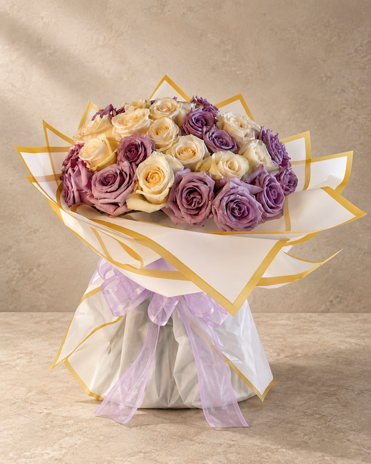 A bouquet of roses wrapped in white paper with gold trim and a sheer lavender ribbon, set against a neutral beige textured background.