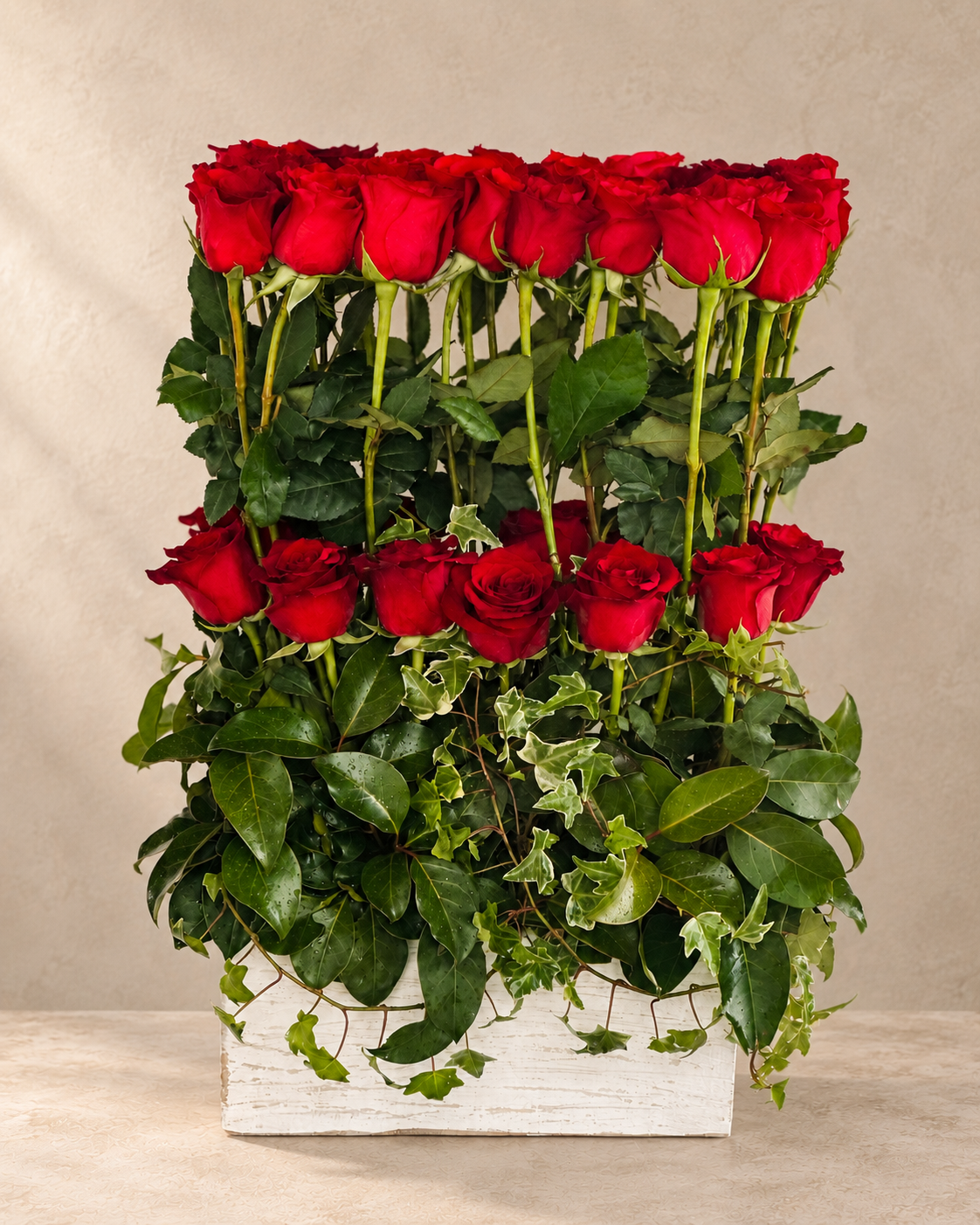 A floral arrangement of several red roses with green leaves and trailing ivy, presented in a white rectangular wooden box against a neutral beige background.