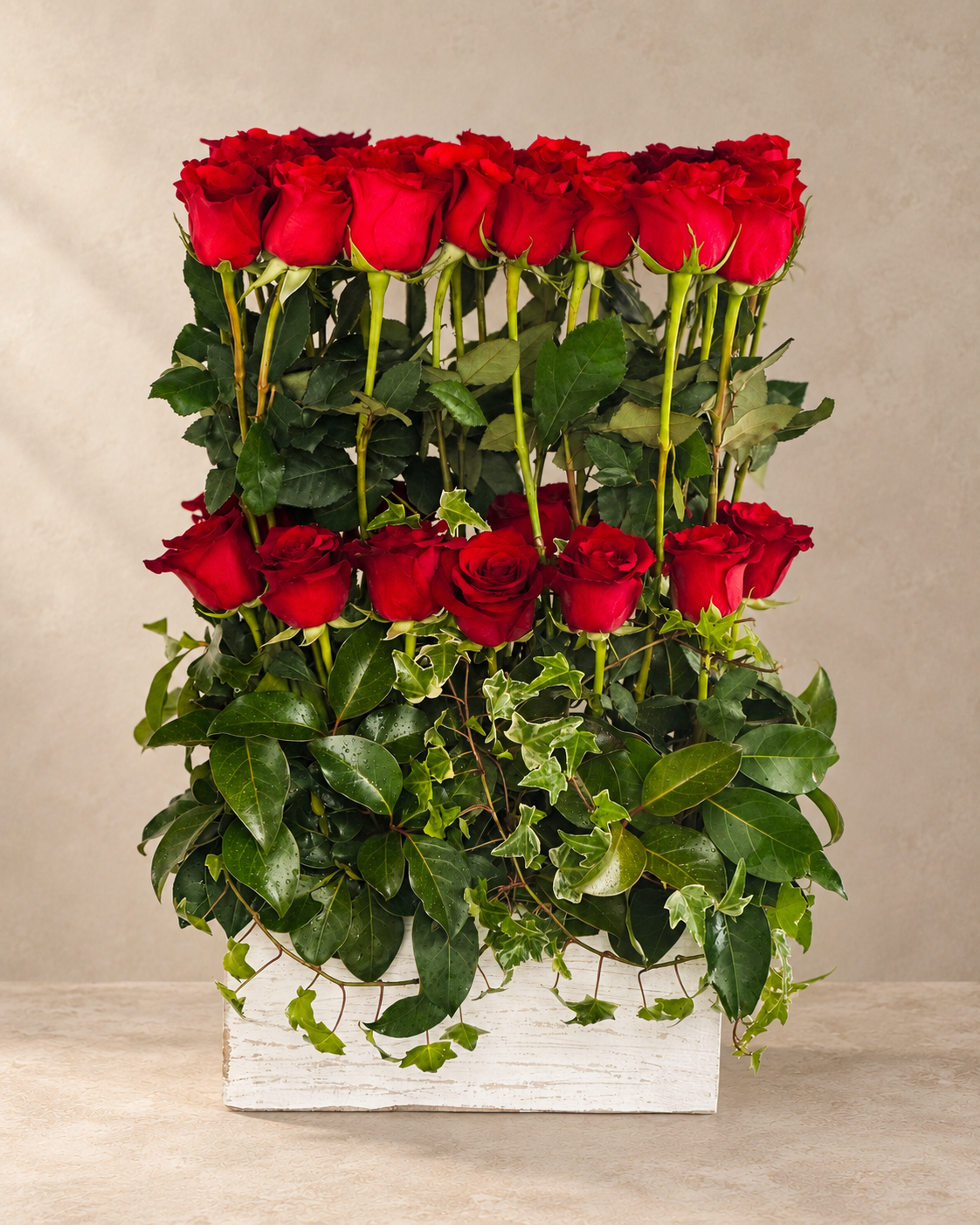 A floral arrangement of several red roses with green leaves and ivy, presented in a white rectangular wooden box against a neutral beige background.