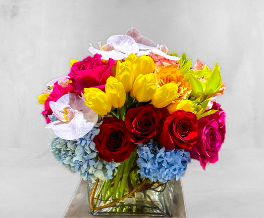 Colorful floral display featuring red and pink roses, yellow tulips, white orchids, and blue hydrangeas in a clear glass vase, highlighting VIP Floristry's premium flower arrangements for all occasions in Houston.