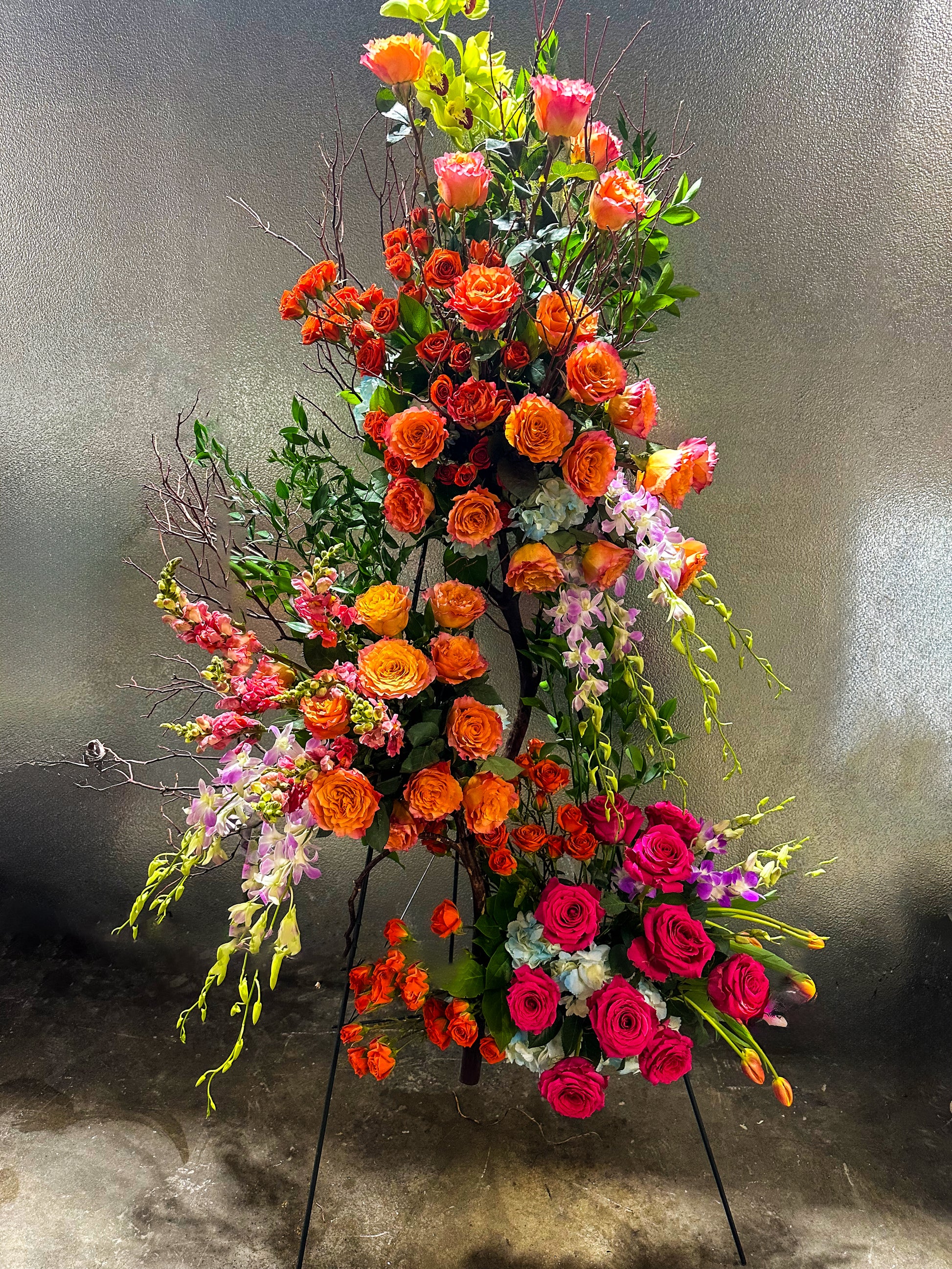 Tall, sophisticated arrangement featuring an array of orange and pink roses, orchids, and natural branches, showcasing VIP Floristry's premium floral artistry for upscale events and weddings in Houston.