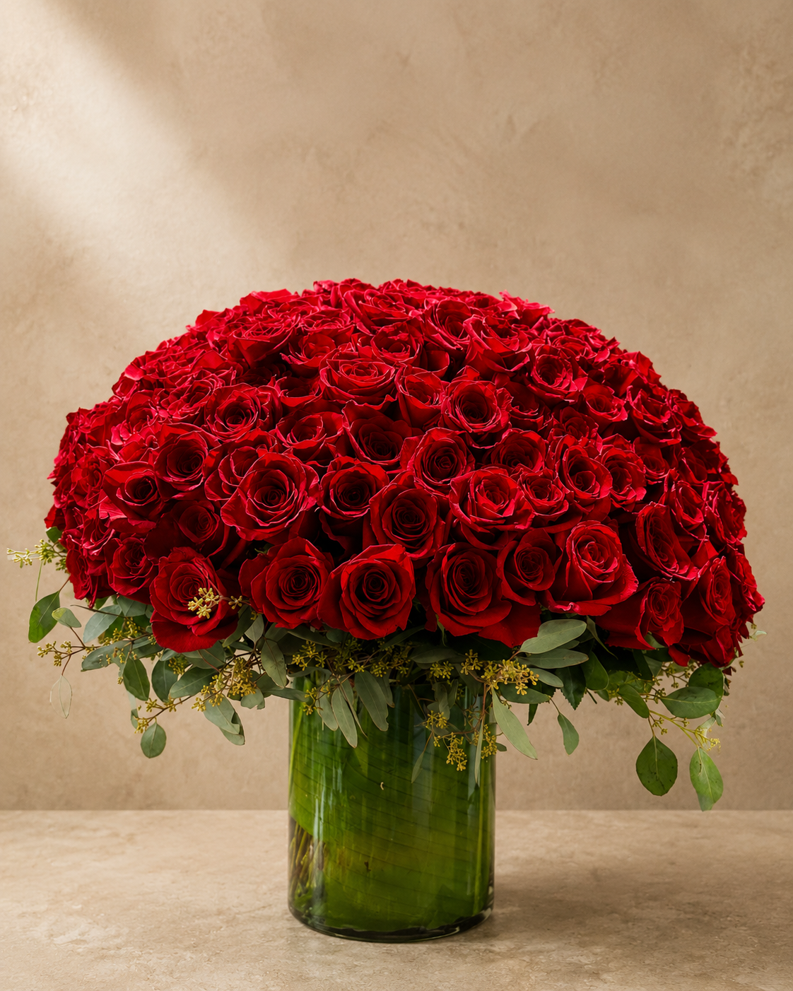 Large dome-shaped arrangement of red roses and eucalyptus in a tall green glass vase against a warm limestone backdrop.