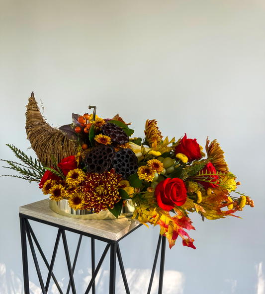 A luxurious fall-themed floral arrangement featuring red roses, orange chrysanthemums, lotus pods, and seasonal accents arranged in a cornucopia display. Perfect for Thanksgiving and autumn celebrations, showcasing the artistry of VIP Floristry in Houston.