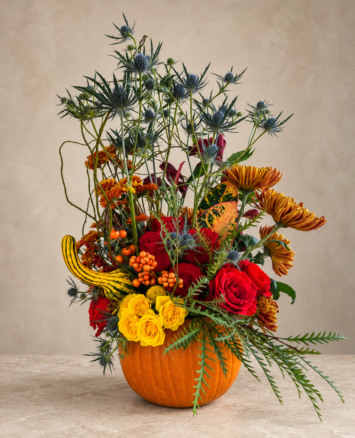 Alternate view featuring blue thistle, red and yellow roses, bronze chrysanthemums, orange berries and a striped ornamental gourd in a pumpkin vessel.