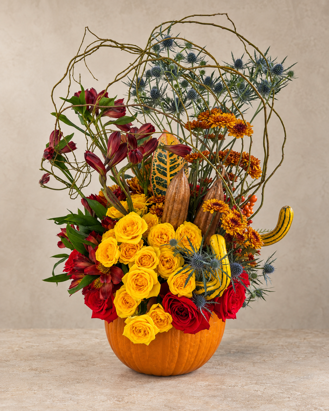 Front view of yellow and red roses, dried pods, striped gourds, blue thistle, autumn chrysanthemums and arching branches arranged in an orange pumpkin vessel.