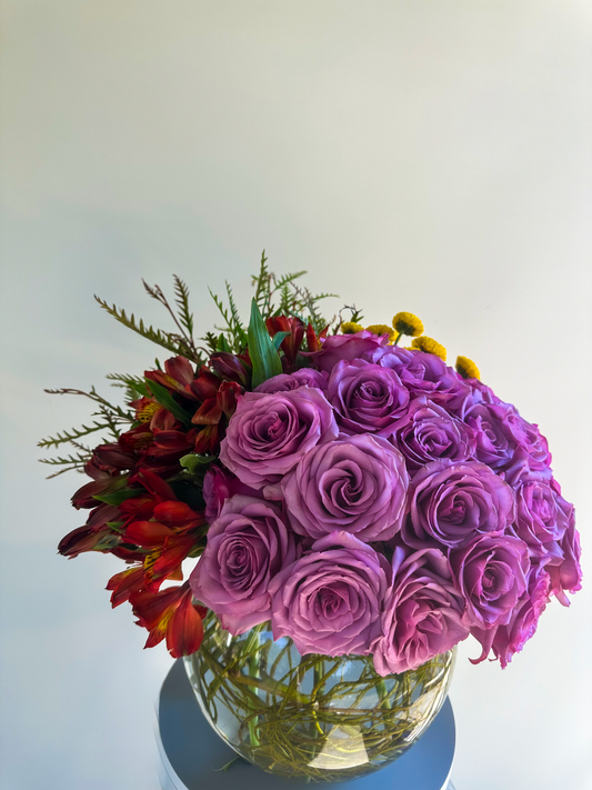 A beautiful arrangement featuring a blend of soft lavender roses, yellow chrysanthemums, and lush greenery in a clear glass bowl. The roses are tightly arranged, adding a sense of opulence, while the greenery provides a natural, earthy touch. This luxury floral arrangement is ideal for Houston events, embodying elegance and sophistication.