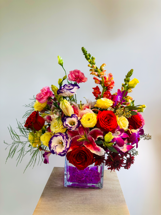 Bright and elegant floral display with red and yellow roses, pink lilies, and purple lisianthus, arranged in a square vase with pink accents, ideal for Houston flower delivery.