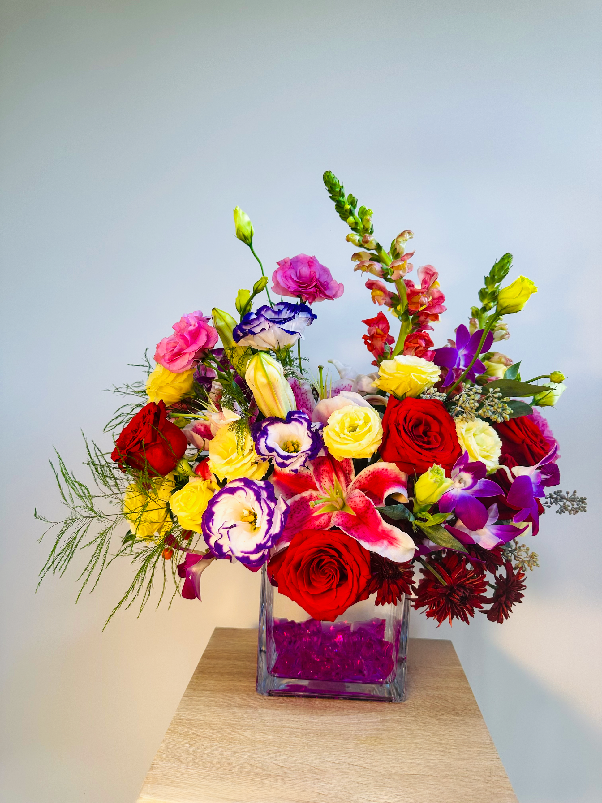 Colorful Houston flower arrangement in a square glass vase with pink gemstones, showcasing roses, lilies, snapdragons, and lisianthus in bold red, yellow, and purple hues.