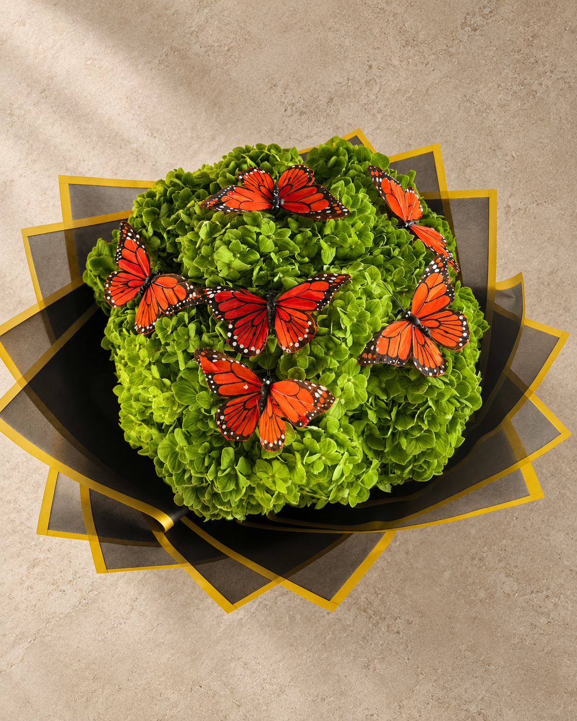 A decorative floral arrangement of bright green hydrangea-like blooms topped with several orange-and-black butterfly decorations, presented in a black and gold geometric tray on a beige textured background.
