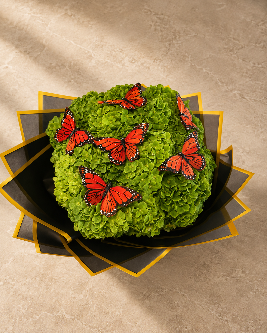 A decorative floral arrangement featuring a rounded mound of bright green hydrangea-like blooms topped with several orange monarch butterflies, presented in a black and gold geometric container on a beige stone surface.