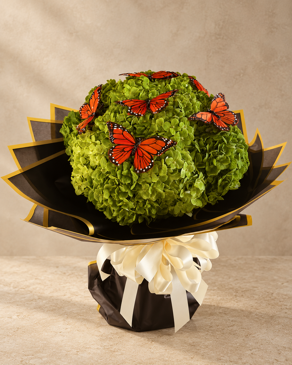 A decorative floral arrangement featuring a round mound of bright green hydrangea-like blooms topped with several orange-and-black butterfly decorations, wrapped in black paper with gold edging and tied with a cream ribbon, set against a warm beige studio background.