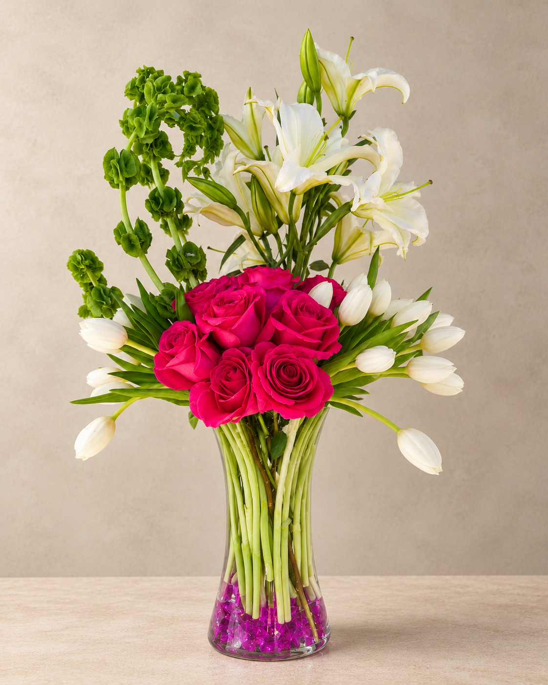 A floral arrangement featuring bright pink roses, white lilies, white tulips, and green foliage in a clear glass vase filled with purple stones, set against a neutral beige background.