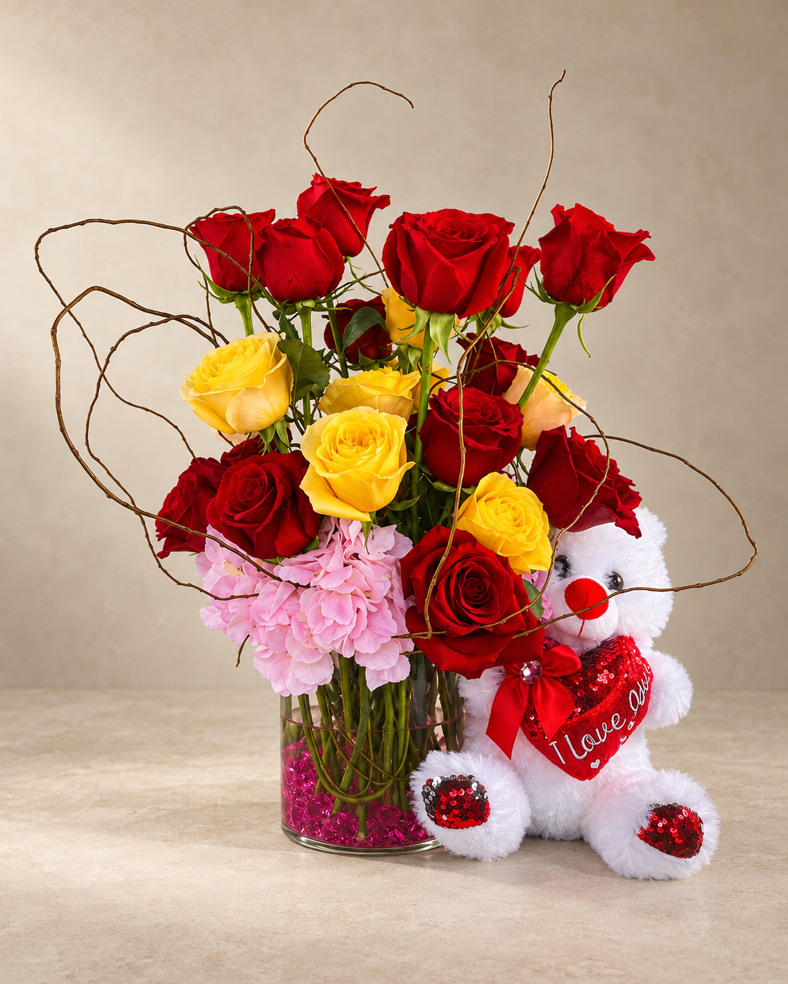 Full studio view of red and yellow roses with pink hydrangea and looping branches in a clear vase with pink stones, accompanied by a white teddy bear holding an “I love you” heart.