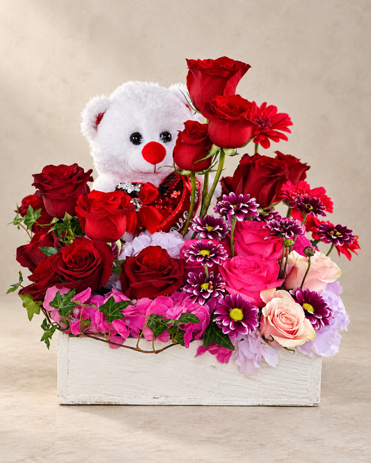 Valentine’s arrangement with a white teddy bear holding a red sequin heart, surrounded by red and pink roses, hydrangea and purple daisies in a white wooden planter.
