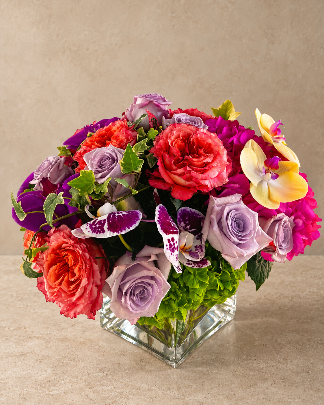 A vibrant floral arrangement featuring red and pink roses, purple and lavender roses, yellow and purple orchids, and green foliage, presented in a clear square glass vase on a beige surface against a textured beige background.