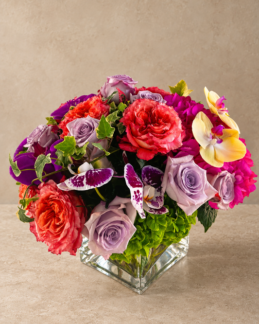 A vibrant floral arrangement featuring red and pink roses, purple and lavender roses, yellow and purple orchids, and green foliage, presented in a clear square glass vase on a beige surface against a textured beige background.