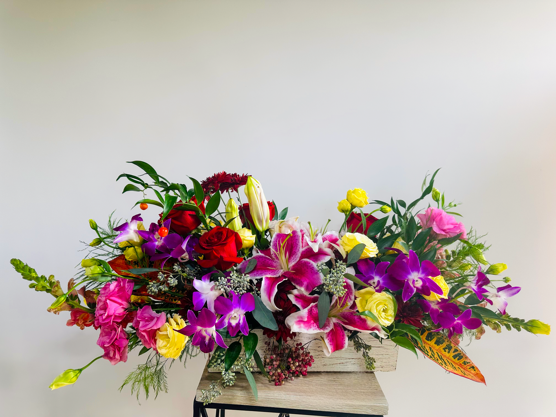 Vibrant floral arrangement with red roses, purple orchids, yellow spray roses, pink lisianthus, and stargazer lilies, designed in a rustic rectangular container. Perfect for Houston floral decor.