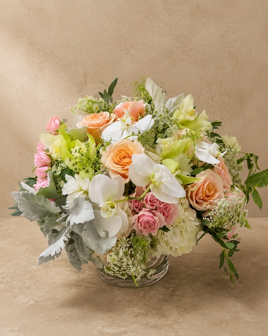 Charming Peach arrangement of peach and blush roses, white orchids, hydrangeas and silvery foliage in a clear glass vase.