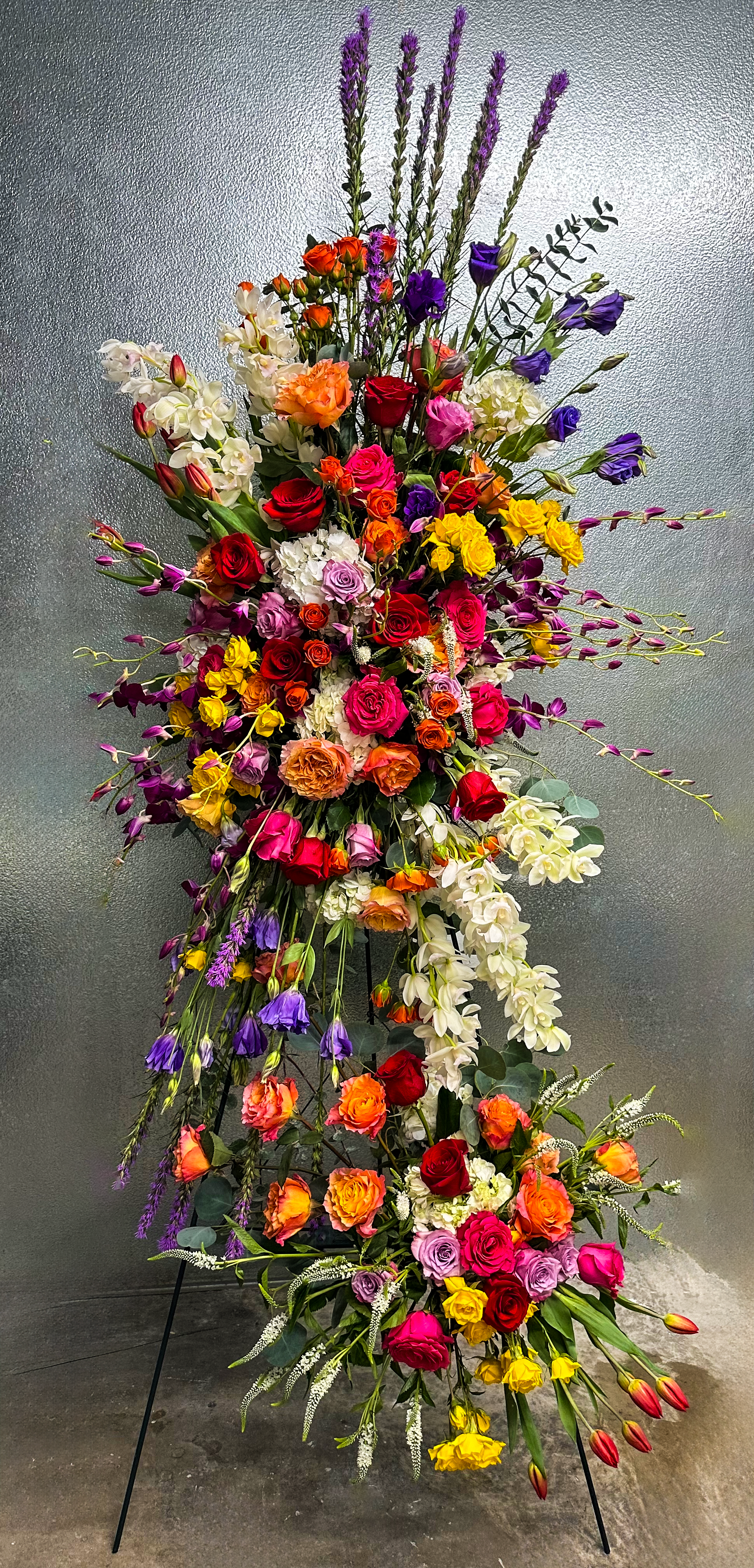 A striking, tall floral arrangement featuring a vibrant mix of roses, hydrangeas, and lisianthus in a spectrum of colors—purple, orange, pink, red, and yellow. This grand design is adorned with elegant cascading orchids and lush greenery, creating a dramatic display suitable for celebrations, memorials, or high-impact events in Houston.