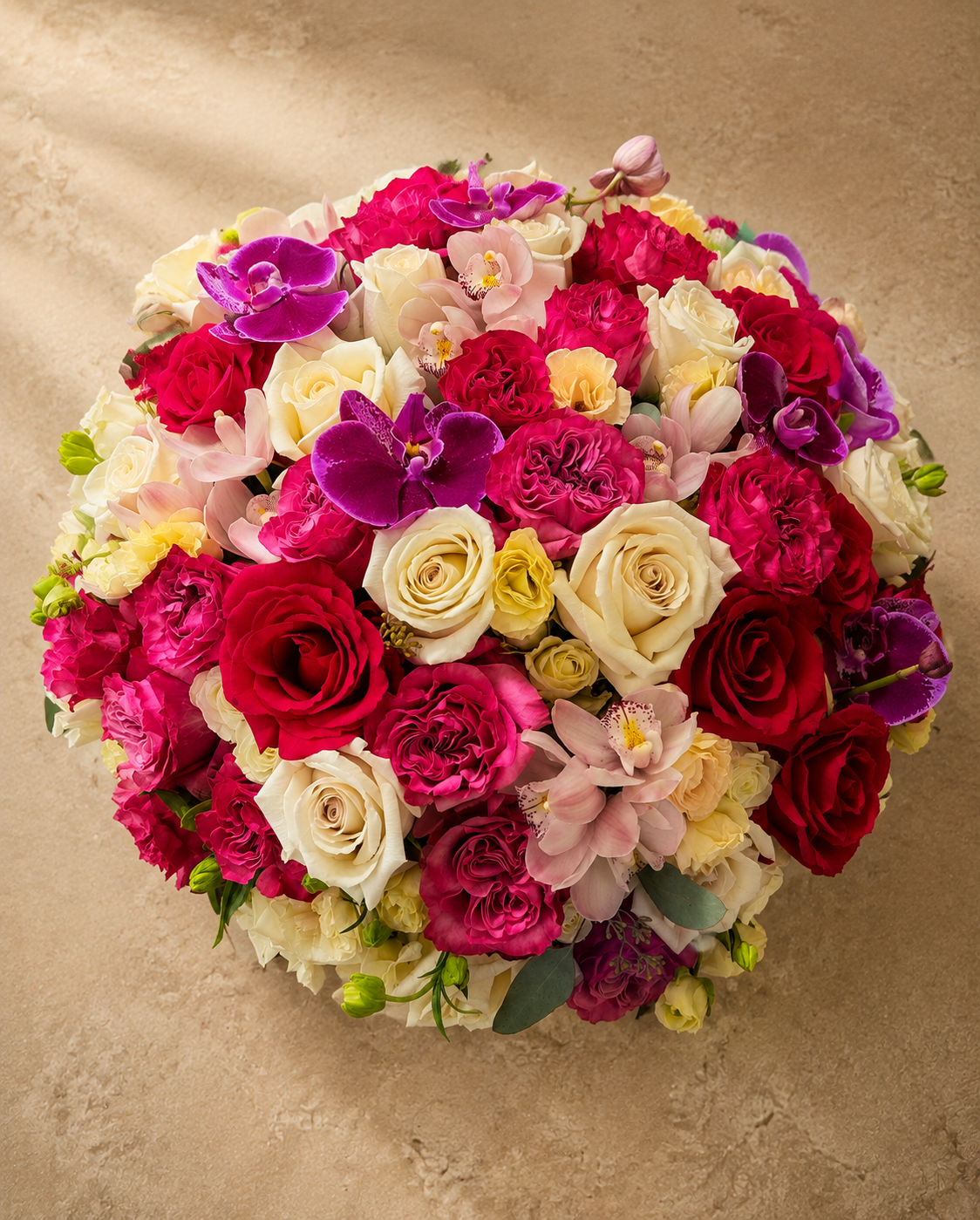 Overhead view of the Jubilation Pavé arrangemen; a large, vibrant round floral arrangement featuring roses, orchids, and carnations in shades of pink, red, purple, cream, and yellow, set against a textured beige background.