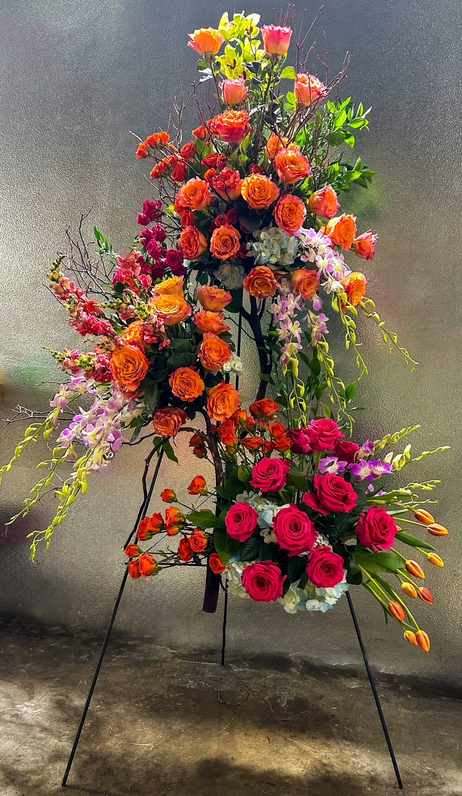 Vibrant floral arrangement with orange and pink roses, orchids, and lush greenery, elegantly cascading on a display stand, creating a stunning visual impact. Perfect for events, this arrangement exemplifies VIP Floristry's luxury floral design expertise in Houston.