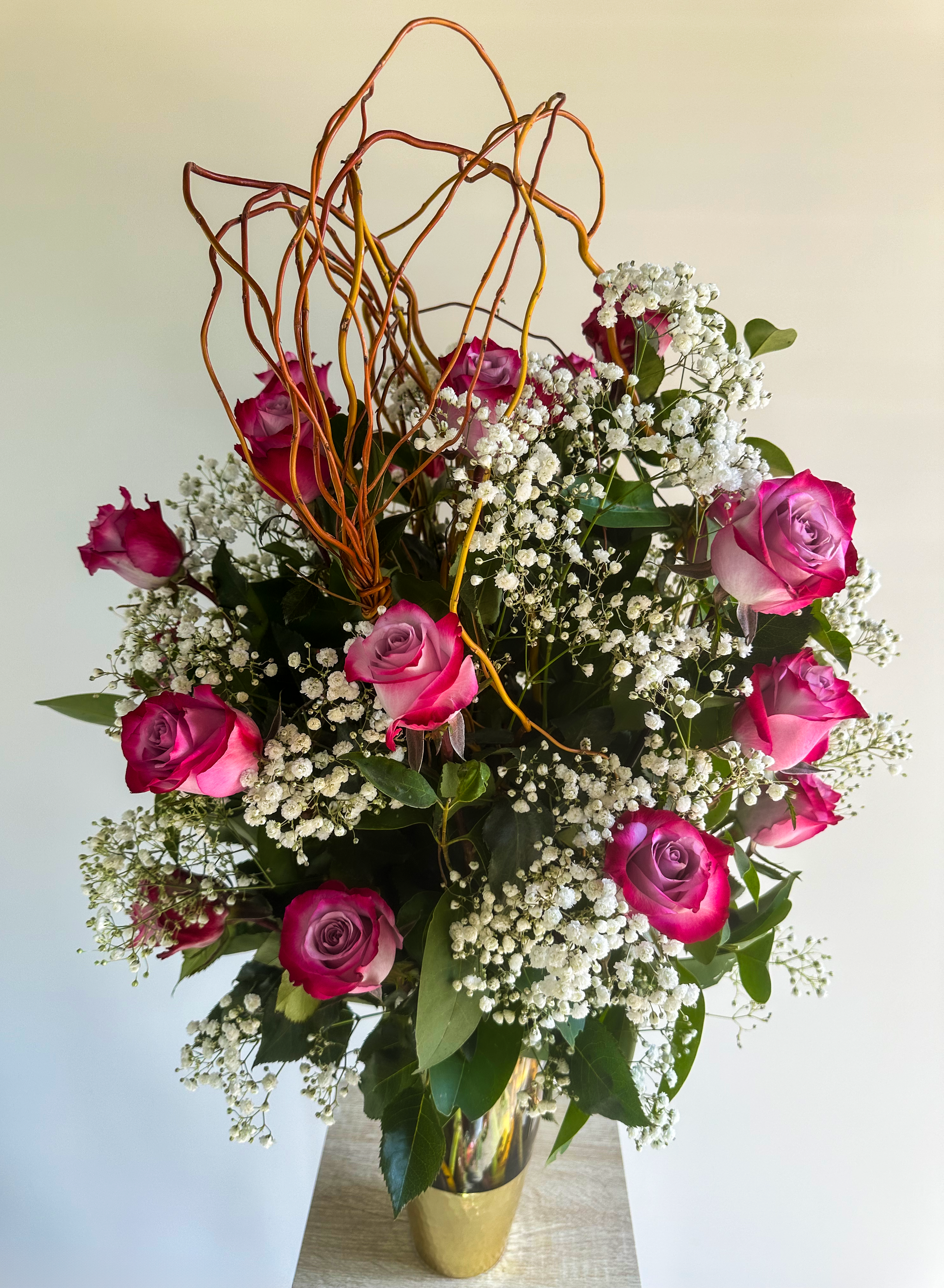 Elegant arrangement of hot pink roses, white baby's breath, and curly willow branches in a gold vase - VIP Floristry, Houston