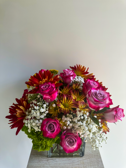 Vibrant floral arrangement with hot pink roses, red and yellow chrysanthemums, white baby's breath, and green accents in a glass vase - VIP Floristry, Houston