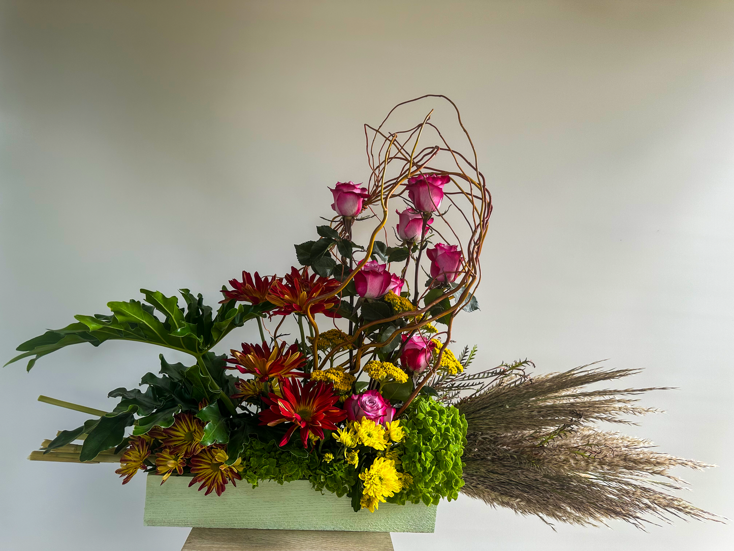 Modern floral arrangement with pink roses, red and yellow chrysanthemums, lush greenery, and textured elements in a rectangular container - VIP Floristry, Houston