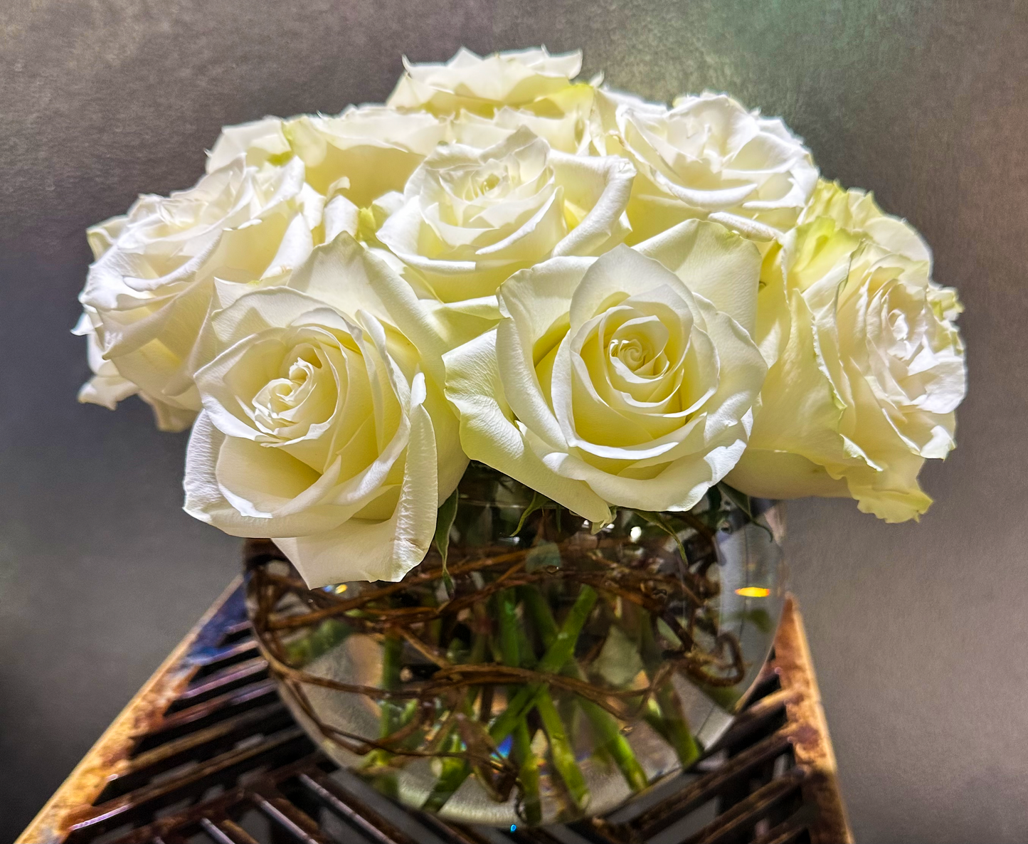 A sophisticated arrangement of pristine white roses in a clear, rounded glass vase. The roses are beautifully clustered, showcasing their delicate petals and rich texture. Twisted willow branches at the base add a natural, rustic touch to the modern design.