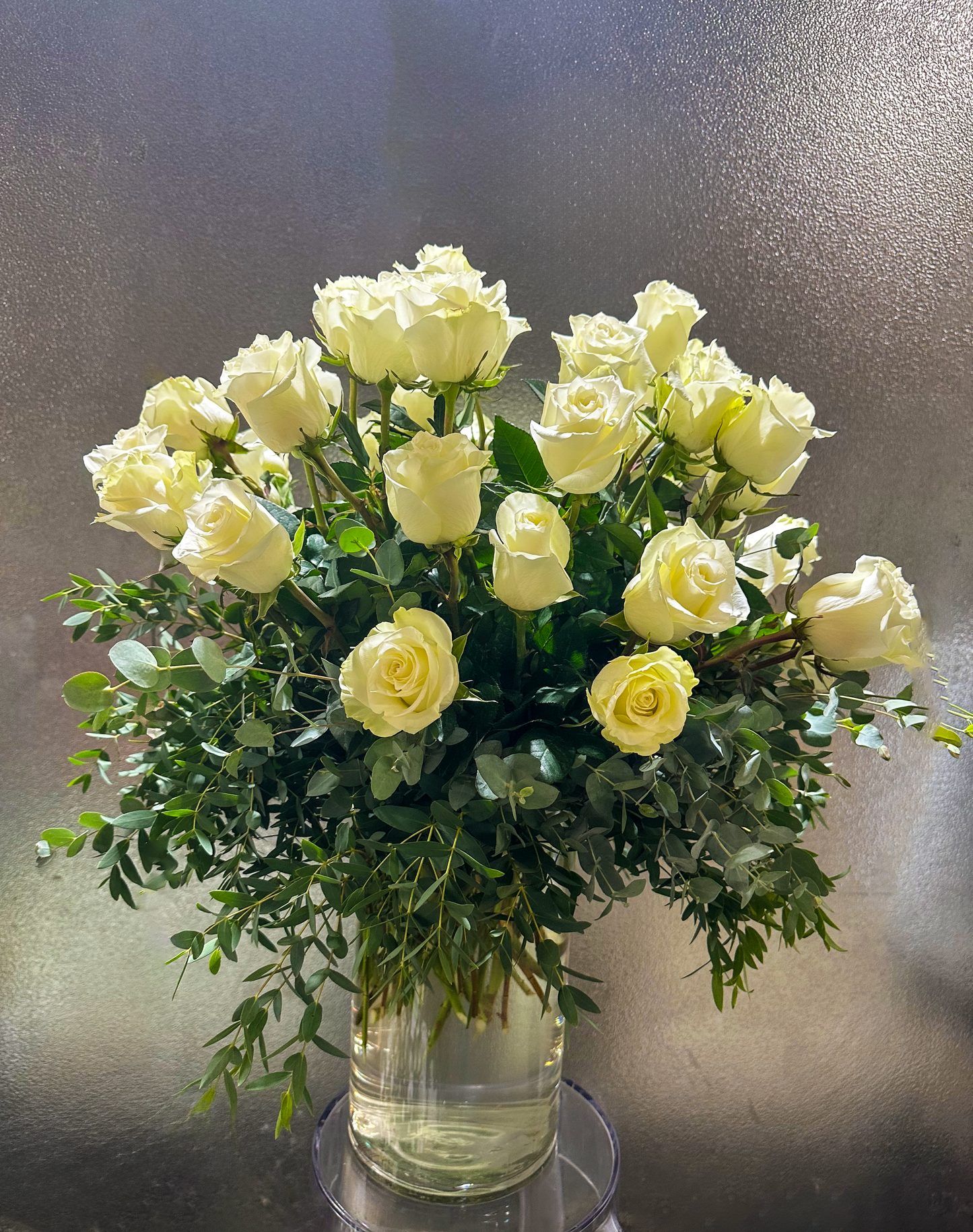 An elegant display of classic white roses arranged in a tall, clear vase, surrounded by lush greenery and eucalyptus, creating a sophisticated and natural look. This arrangement brings elegance and a touch of nature to any Houston home or event, making it a fitting choice for those seeking premium floral arrangements in the area.