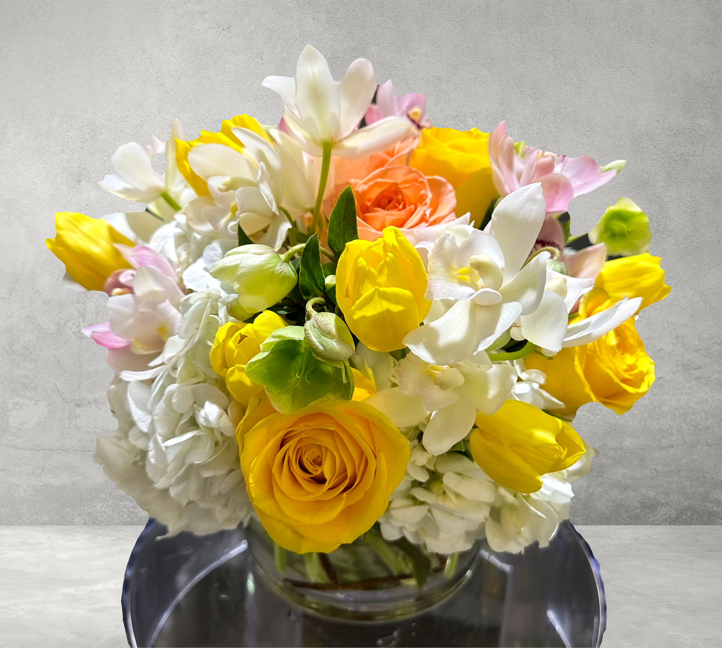 A vibrant floral arrangement featuring a blend of yellow tulips, white orchids, peach roses, and soft pink accents in a clear glass vase. This cheerful bouquet exudes a lively energy, perfect for brightening any space in Houston.